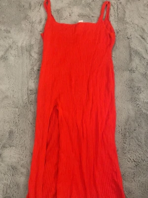 Röd ribbad långklänning med slits - Säljer en snygg röd långklänning med ribbad struktur och smala axelband. Klänningen har en slits på sidan som ger en cool touch. Perfekt för dig som gillar färgstarka och stilrena plagg med lite edge. Kan eventuellt tänka mig gå ner i pris