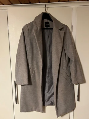 Grå kappa från New Look Petite - Grä kappa från New Look Petite med klassisk krage och två fickor framtill. Rak passform och stilren design som funkar till många olika outfits. Perfekt för dig som gillar enkel och clean stil. Fodrad insida och i mycket bra skick. 