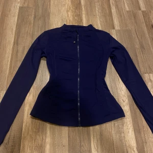 Mörkblå träningsjacka  - Snygg mörkblå träningsjacka med figurnära passform och hel dragkedja framtill. Jackan har lång ärm, hög krage och ett litet guldfärgat logodetalj vid halsen. Perfekt för träning.