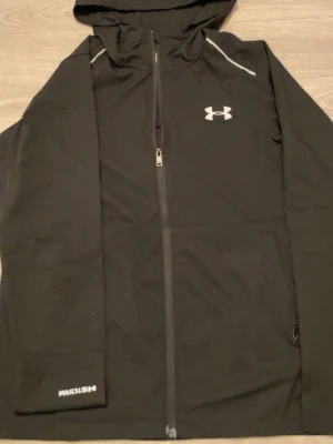 Svart vindjacka från Under Armour - Svart vindjacka från Under Armour med huva och hel dragkedja. Jackan har vita loggor på bröstet, ärmen och ryggen samt reflekterande detaljer vid axlarna. Tillverkad i lätt polyester med meshfoder för extra komfort. Perfekt för träning utomhus.