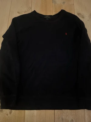 Svart sweatshirt från Polo Ralph Lauren - Klassisk svart sweatshirt från Polo Ralph Lauren med rund halsringning och den ikoniska röda logobrodyr på bröstet. Tröjan har lång ärm och är tillverkad i mjuk bomull för en skön och avslappnad look. Använd men har inga defekter eller hål. Fraktar på 1-2 dagar!