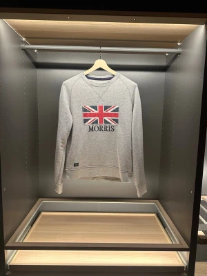Grå sweatshirt från Morris  - Grå sweatshirt från Morris i bomull med storbritaniens flagga och Morris-tryck på bröstet. Klassisk rund halsringning och långa ärmar. Perfekt för dig som gillar stilren design med brittisk touch. Det är storleken S men skulle säga att den passar mer som en M. Hör av er vid frågor och funderingar.