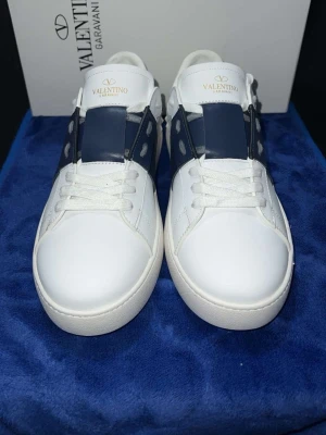 Valentino Garavani Open sneakers vit/blå - Snygga Valentino Garavani Open sneakers i vitt skinn med bred marinblå läderdetalj över mitten och vita snören. Rund tå och platt sula med mönstrad undersida. Ikonisk logga på plösen och coola nitar på sidorna. Perfekt för dig som gillar exklusiv streetstyle.