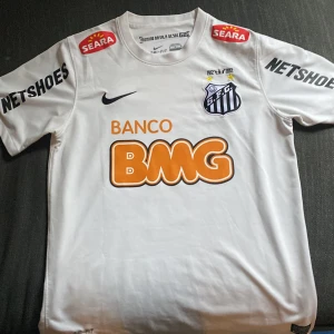 Neymar Jr Santos tröja 2012 - Bilderna visar en vit fotbollströja från Nike med Santos FC-logga, sponsortryck och Neymar Jr samt nummer 11 på ryggen. Tröjan är i storlek M och tillverkad i Brasilien med Dri-Fit-material.