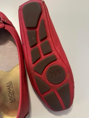 Röda ballerinas från Michael Kors - Snygga röda ballerinas från Michael Kors med guldfärgad logga framtill. Skorna är i skinn med rund tå och platt sula. Insidan har en guldfärgad detalj och yttersulan har bruna gummipartier för extra grepp. Perfekta för dig som vill ha en klassisk men trendig look.