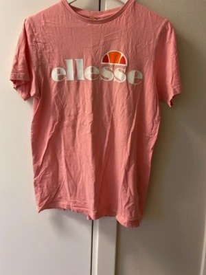 Rosa t-shirt från Ellesse - Säljer en rosa t-shirt från Ellesse med stort vitt tryck och logga på bröstet. T-shirten har rund hals och korta ärmar, tillverkad i mjuk bomull. Perfekt för dig som gillar sportiga och snygga streetwear-plagg.