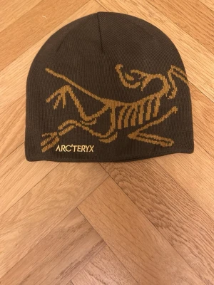 Brun mössa från Arc'teryx - Säljer en brun mössa från Arc'teryx med ett stort guldfärgat fossiltryck på framsidan och logga broderad i gult. Mössan är stickad och har en klassisk, rundad form som sitter skönt på huvudet. Perfekt för kalla dagar.