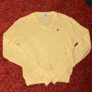 Gul v-ringad tröja från Von Cort - Säljer en ljusgul v-ringad tröja från Von Cort med ett broderat grodmärke på bröstet. Tröjan är långärmad och har ribbade muddar vid ärmslut och nederkant. Materialet är 100% akryl och den har en mjuk, stickad känsla. Perfekt för dig som gillar unika detaljer.