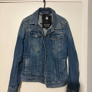 Blå jeansjacka från G-Star RAW - Säljer en klassisk blå jeansjacka från G-Star RAW i storlek S. Jackan har knappar framtill, två bröstfickor med lock och knapp samt sidofickor. Snygg krage och långärmad modell, perfekt för att ge din outfit en cool touch.