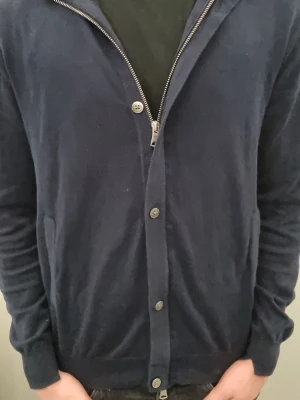 Marinblå kofta från J.Lindeberg - Snygg marinblå kofta från J.Lindeberg med både dragkedja och knappar framtill. Tröjan har lång ärm och är tillverkad i ett mjukt stickat material som känns skönt mot huden. Perfekt för lager-på-lager och chill stil.