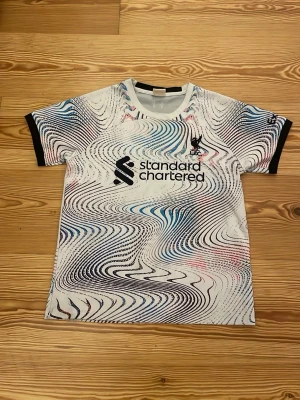 Liverpool Salah tröja med mönster strl 164 - Säljer en Liverpool FC matchtröja med M. Salah och nummer 11 på ryggen. Tröjan har ett unikt psykedeliskt mönster i svart, blått och rosa på vit botten, med svarta detaljer vid ärmslut och krage. Materialet är polyester och trycket är i svart.