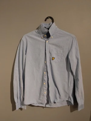 Ljusblå skjorta från Lyle & Scott - Snygg ljusblå skjorta från Lyle & Scott med klassisk krage och knappar framtill. På bröstfickan sitter den ikoniska gula fågeln som broderad detalj. Skjortan är långärmad och gjord i mjuk bomull, perfekt för en chill och stilren look.
