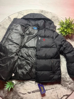 Svart pufferjacka från Polo Ralph Lauren - Svart pufferjacka från Polo Ralph Lauren med huva, dragkedja och flera fickor. Jackan har ett rött broderat Polo-logo på bröstet och quiltad design. Materialet är syntet och dun, perfekt för kalla dagar. Snygg och stilren look med normal passform.