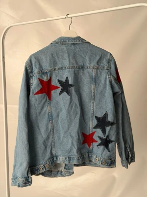 Jeansjacka med stjärnor  - Kollektion: Utility                                                 Jeansjacka med stora röda och mörkblå stjärnor på ryggen och ärmen. Jackan har en loose passform och är perfekt för dig som vill sticka ut med coola detaljer.