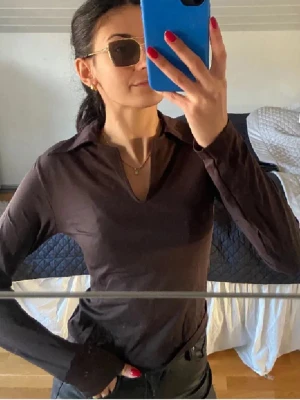 Brun långärmad polotopp - Säljer en brun långärmad polotopp med krage och v-ringning. Toppen har en slim passform och är tillverkad i ett mjukt material som sitter snyggt på kroppen. Perfekt att styla med jeans eller kjol för en trendig look.