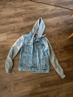 Blå jeansjacka med grå hoodie - Säljer en snygg jeansjacka i ljusblå denim med gråa sweatshirt-ärmar och huva. Hqr haft ett hål som nu är ihopsydd 