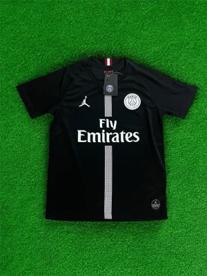 PSG fotbollströja  - Bilden visar ingen sko, utan en svart PSG fotbollströja med Fly Emirates-tryck och Jordan-logga. Ingen information om skor finns.