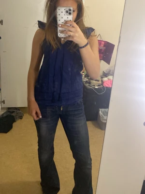 Mörkblå blus med volang från Max - Säljer en mörkblå blus från Max med snygga volangdetaljer vid axlarna och broderade paneler framtill. Blusen är ärmlös och har en lätt v-ringad hals. Materialet är mjukt och känns som bomull, perfekt för vår och sommar.