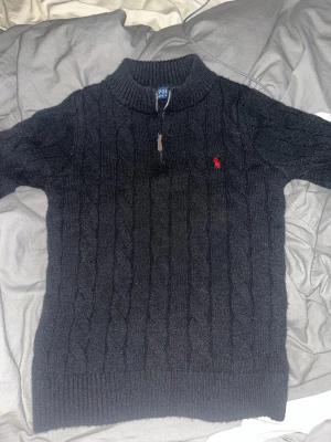 Svart stickad half zip tröja Polo Ralph Lauren - Svart kabelstickad tröja från Polo Ralph Lauren med half zip och hög krage. Klassisk röd logga broderad på bröstet. Tröjan är långärmad och har ribbade muddar vid ärmslut och nederkant. Perfekt för dig som gillar stilrena och tidlösa plagg.