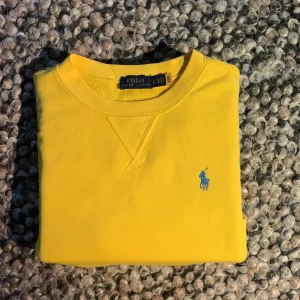 Gul sweatshirt från Polo Ralph Lauren - Säljer en gul sweatshirt från Polo Ralph Lauren i storlek S. Tröjan har ett blått broderat Polo-logo på bröstet, rund hals och långa ärmar. Materialet är mjuk bomull och passformen är normal. Perfekt för dig som gillar färgstarka plagg och klassisk stil.