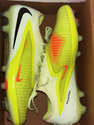 Nike Phantom fotbollsskor gul/svart - Nike Phantom fotbollsskor i en fräsch mix av ljusgul, neon och svart med orange detaljer. Skorna har snörning, platt sula med dobbar och är tillverkade i syntetmaterial med stickad ovandel för extra komfort och grepp. Perfekta för dig som vill sticka ut på planen.