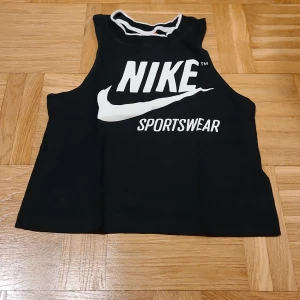 Svart linnetopp från Nike XS - Svart linnetopp från Nike i storlek XS med vit Nike Sportswear-logga framtill. Toppen är ärmlös och har en vit kant runt halsen. Perfekt för sportiga vibes och chill dagar. Materialet är mjuk bomull som känns skönt mot huden.
