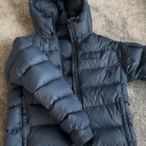 Mörkblå Peak Performance dunjacka - Säljer en mörkblå dunjacka från Peak Performance, modell M Frost DJ. Jackan har en stor huva, dragkedja framtill och breda, quiltade sektioner. Perfekt för kalla dagar och har en clean, sportig look. Materialet är polyester och dun. Storlek small