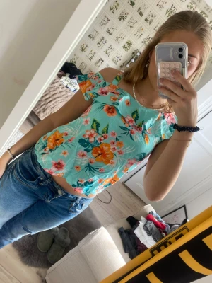 Blommig offshoulder topp från H&M - Säljer en superfin blommig topp från H&M i turkos med färgglada blommor i orange, rosa och grönt. Toppen har offshoulder-modell med volangdetalj och smala axelband. Perfekt för sommarens alla tillfällen!