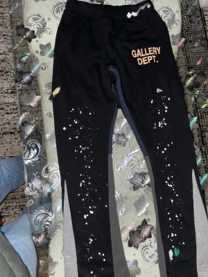 Svarta mjukisbyxor Gallery Dept. - Säljer ett par svarta mjukisbyxor från Gallery Dept. med elastisk midja och coola färgstänk i vitt, gult och grönt. Byxorna har tryckt logga på ena låret och kontrasterande grå paneler längs benen. Perfekta för en avslappnad och trendig look.