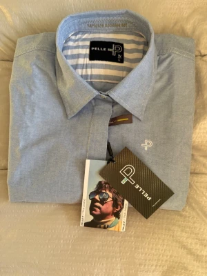 Blå skjorta från Pelle P i bomull - Snygg ljusblå skjorta från Pelle P i bomull. Klassisk krage, långa ärmar med knappar vid manschetten och diskret broderad logga på bröstet. Insidan av kragen har blåvita ränder för en extra detalj. Perfekt för en clean och stilren look. Spliter ny dessutom och i storlek L. 