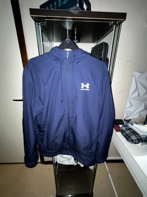 Blå vindjacka från Under Armour - Snygg marinblå vindjacka från Under Armour med huva och dragkedja framtill. Jackan har meshfoder på insidan, två sidofickor och logga i vitt på bröstet. Perfekt för dig som gillar sportig stil och vill ha något lätt och funktionellt.trasig dragkedja 