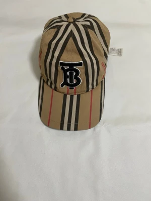 Randig Burberry keps i bomull - Säljer en snygg keps från Burberry med klassiskt randigt mönster i beige, svart, vit och röd. Kepsen har en broderad TB-logga framtill och är tillverkad i 100% bomull. Perfekt accessoar för att lyfta din stil.