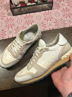 Valentino Rockrunner sneakers beige/vit - Säljer ett par Valentino Rockrunner sneakers i beige och vitt med coola nitar på hälen. Skorna har en mix av mocka och skinn, samt kamouflagemönster på ovandelen. Snygg kontrast med svart sula och klassisk snörning. Perfekta för dig som gillar exklusiva och trendiga sneakers. Lite stora på mig i bredden men passar i längden så skulle säga dom passar i storlek 44-45 