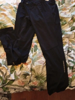 Svarta täckbyxor med reflexdetalj - Svarta täckbyxor med elastisk midja och rak passform. Byxorna har en diskret reflexdetalj längs ena benet och är gjorda i ett slitstarkt syntetmaterial som passar perfekt för medium kalla dagar. Snygga att ha över vanliga kläder när du ska ut och röra på dig. Varumärket är PRO TOUCH 