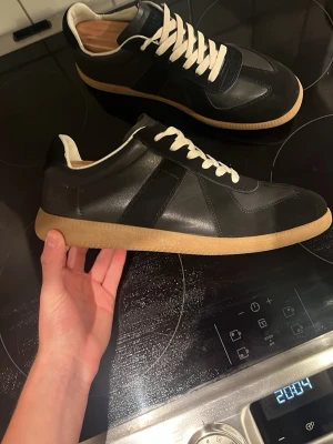 Maison Margiela skor  - Maison Margiela sneakers i svart skinn och mocka med vita snören och beige gummisula. Klassisk rund tå och platt sula, med diskret logotyp på tungan och undersidan. Snygg retrostil som funkar till många outfits.