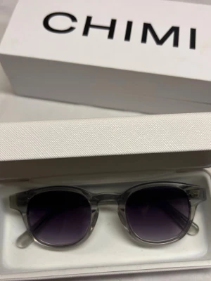 Chimi - Bilderna visar ett par solglasögon från CHIMI med transparent båge och mörka glas, samt tillhörande förpackning. 8/10 skick använd en sommar.