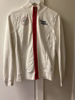 Vit ziptröja från Tommy Hilfiger - Säljer en vit långärmad ziptröja från Tommy Hilfiger med röd dragkedja och broderad logga på bröstet. Tröjan har ståkrage, två fickor och ett snyggt märke på vänster sida. Perfekt för dig som gillar klassisk streetstil.
