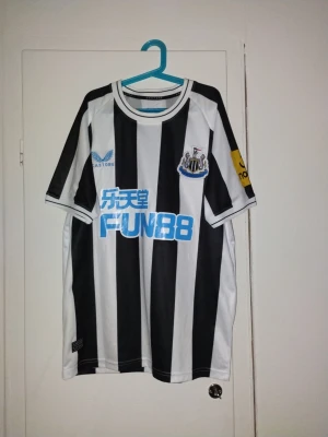 Newcastle United Trippier matchtröja S - Säljer en Newcastle United matchtröja med Trippier 15 på ryggen. Tröjan är svartvit med breda ränder, blå Castore-logga och klubbmärke på bröstet samt sponsortryck i blått och gult. Tillverkad i polyester med rund hals och korta ärmar.