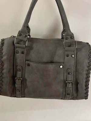 Grå handväska med detaljer - Snygg grå handväska i skinnimitation med både handtag och avtagbar axelrem. Väskan har dekorativa nitar, spännen och flätade sidor. Framtill finns en praktisk ficka och insidan har en annan ficka. Mått: längd: 28cm bredd: 22cm handtag: 15cm (hälften) extra rem: 47cm