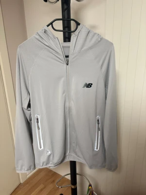 Ljusgrå vindjacka New Balance - Snygg ljusgrå vindjacka från New Balance med huva och hel dragkedja framtill. Jackan har två fickor med dragkedja och reflekterande detaljer. Perfekt för träning eller när du vill ha en sportig look. Materialet är lätt och känns skönt mot huden.