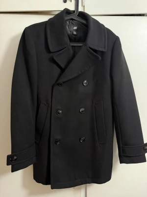 Svart skepparkavaj från H&M - Stilren svart skepparkavaj från H&M med klassisk dubbelknäppning och bred krage. Jackan har två sidofickor och dekorativa knappar vid ärmsluten. Perfekt för dig som vill ha en tidlös look med lite edge.