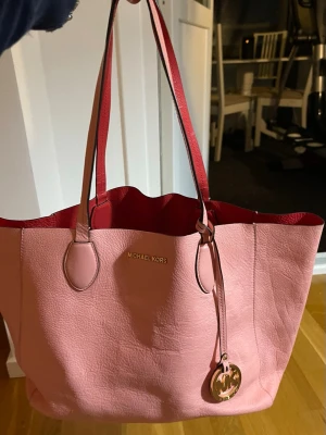 Rosa shoppingväska från Michael Kors - Stor och rymlig shoppingväska från Michael Kors i ljusrosa skinn med röda inslag på insidan och handtag. Väskan har guldfärgad MK-logga och detaljer, samt mjuk struktur. Perfekt för dig som vill ha plats för allt och samtidigt se stylish ut.