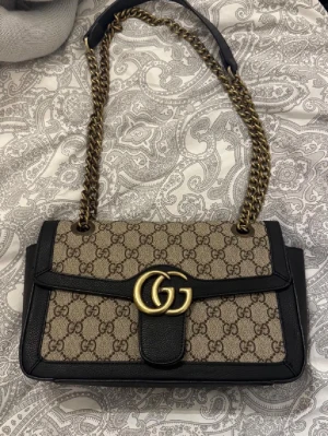 Gucci axelväska med kedja och GG-logga - Säljer en snygg axelväska från Gucci med klassiskt GG-mönster i beige och brunt. Väskan har svarta skinnkanter, guldfärgad kedja och stor GG-logga framtill. Insidan har dragkedja och foder i ljus färg. Perfekt för dig som gillar lyxiga detaljer och ikonisk design.