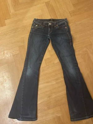 True Religion Joey Low Rise Flare jeans - Säljer ett par mörkblå True Religion Joey Low Rise Flare jeans i storlek 24. De har låg midja, utställda ben och snygga kontrastsömmar i orange. Baksidan har två fickor med lock och dekorativa knappar. Klassisk jeanslook med slitningar och dragkedja framtill.