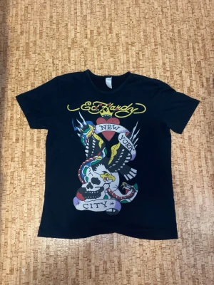 Svart Ed Hardy t-shirt med tryck - Svart t-shirt från Ed Hardy med färgstarka tryck både fram och bak. Framsidan har motiv med hjärta, örn, dödskalle och texten 'New York City'. Baksidan har dödskalle med racingtema och texten 'Los Angeles Dead or Alive'. Klassisk rund hals och korta ärmar. Materialet är mjuk bomull. Några stenar har lossnat.
