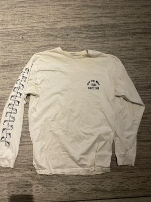 Vit långärmad topp från Vans - Vit långärmad topp från Vans med tryck på bröstet 'Off The Wall Since 1966' och coolt pingvinmotiv på ryggen. Ena ärmen har grafiska tryck i svart. Rund halsringning och avslappnad passform, perfekt för en chill streetwear-look.