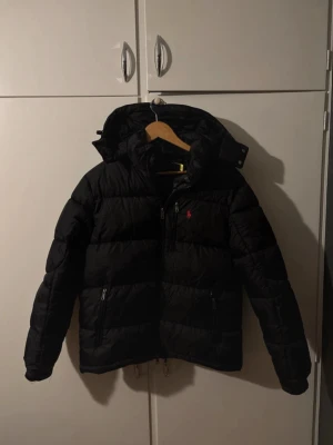 Svart pufferjacka från Polo Ralph Lauren - Säljer en svart pufferjacka från Polo Ralph Lauren med huva och dragkedja framtill. Jackan har det klassiska röda Polo-loggan broderad på bröstet och två fickor med dragkedja. Perfekt för kalla dagar och riktigt snygg streetstil.