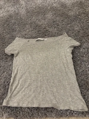 Grå offshoulder topp med spets  - Säljer en ljusgrå offshoulder topp från lager 157 i storlek M. Toppen har korta ärmar och en fin spetsdetalj längs halsringningen. Materialet är mjukt och känns som bomull, perfekt för en chill och stilren look. Helt oanvänd 💕