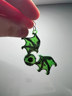 Grön svarta glittriga fladdermus örhängen - Unika örhängen i form av fladdermusvingar med ett öga i mitten. Färgen är intensivt grön med glittriga detaljer och svarta konturer som ger en cool kontrast. designen är riktigt edgy och sticker ut.
