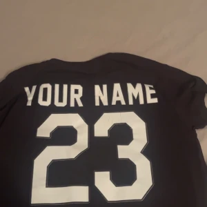 Svart Off-White x Sox t-shirt - Svart t-shirt från Off-White med coola vita tryck framtill och stort tryck med texten 'YOUR NAME' och siffran 23 på ryggen. T-shirten har korta ärmar och en klassisk rund hals. Materialet känns som mjuk bomull, perfekt för en chill streetwear-look.